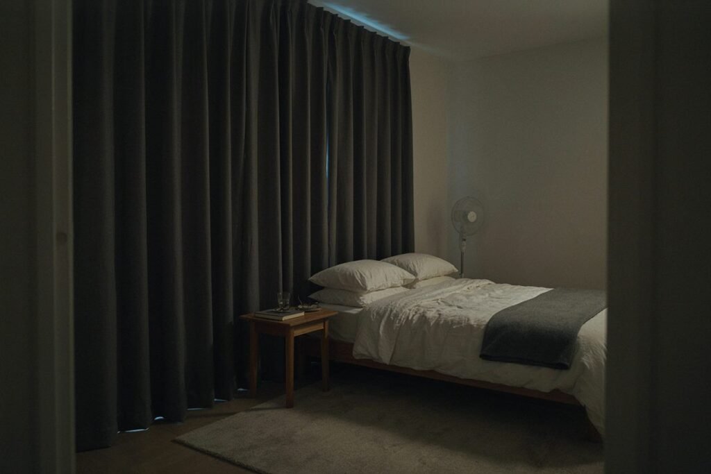 Quarto escuro com cortina blackout fechada, ventilador e cama arrumada